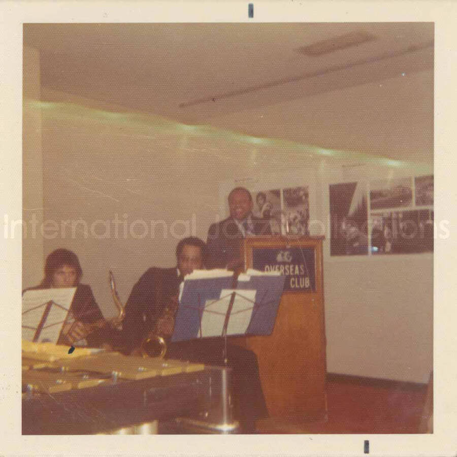 3 1/2 x 3 1/2 inch photograph. Lionel Hampton and band at the Overseas Press Club of America. A banner on the wall reads: Welcome Lionel Hampton, America's Good Will Ambassador of Jazz