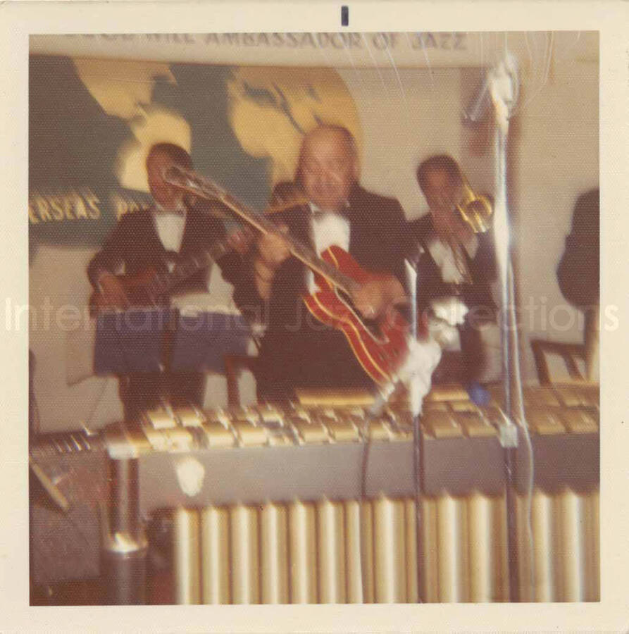 3 1/2 x 3 1/2 inch photograph. Lionel Hampton and band, which includes guitarist Billy Mackel, at the Overseas Press Club of America. A banner on the wall reads: Welcome Lionel Hampton, America's Good Will Ambassador of Jazz
