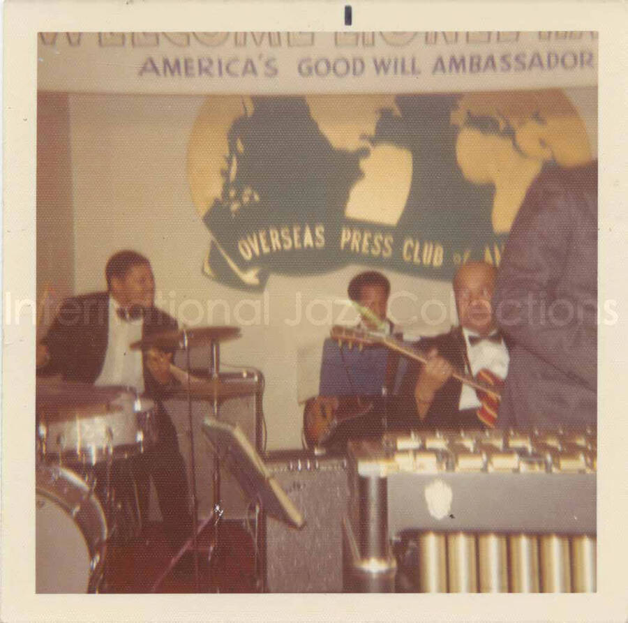 3 1/2 x 3 1/2 inch photograph. Lionel Hampton and band, which includes guitarist Billy Mackel, at the Overseas Press Club of America. A banner on the wall reads: Welcome Lionel Hampton, America's Good Will Ambassador of Jazz