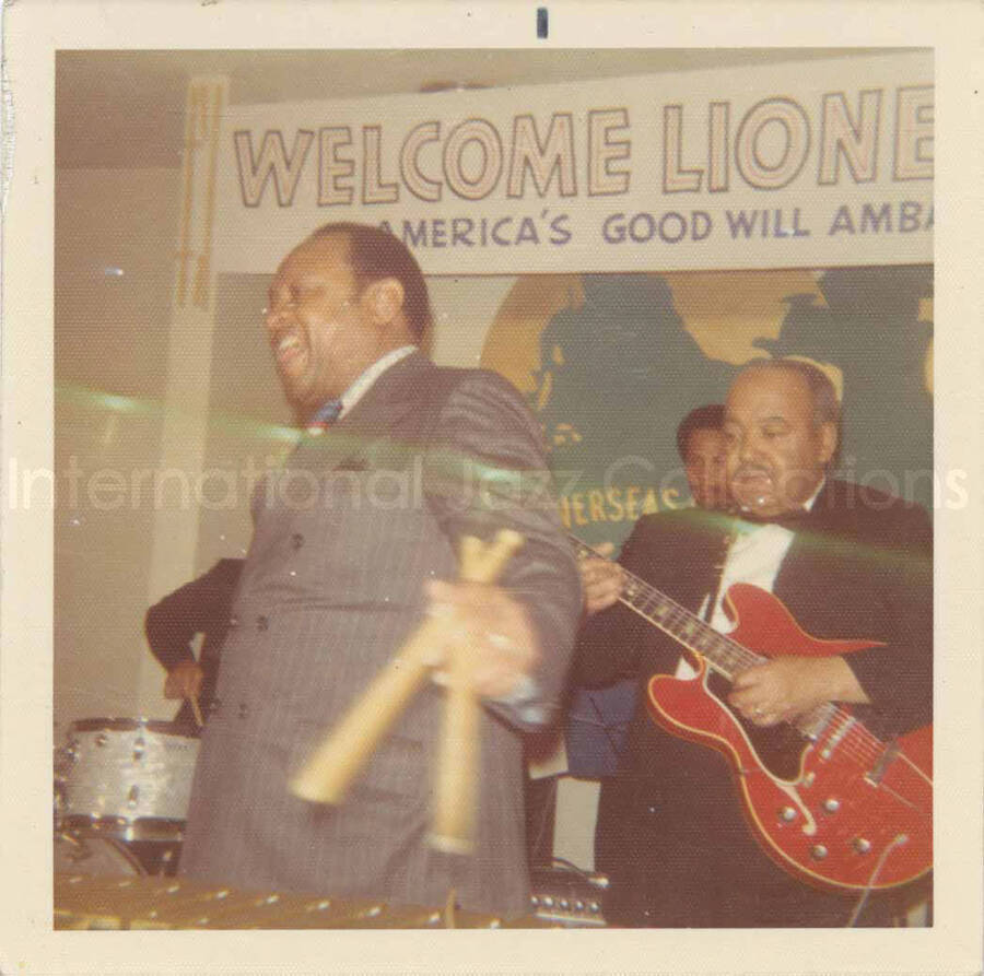 3 1/2 x 3 1/2 inch photograph. Lionel Hampton and band, which includes guitarist Billy Mackel, at the Overseas Press Club of America. A banner on the wall reads: Welcome Lionel Hampton, America's Good Will Ambassador of Jazz