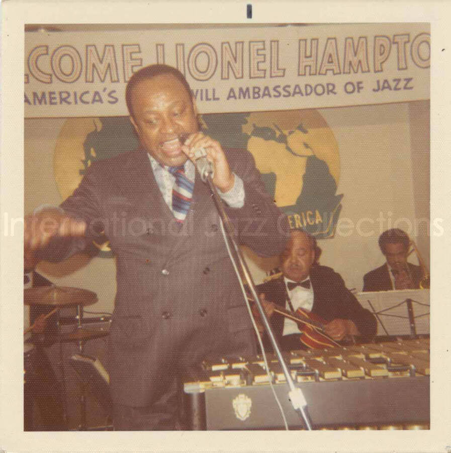 3 1/2 x 3 1/2 inch photograph. Lionel Hampton and band, which includes guitarist Billy Mackel, at the Overseas Press Club of America. A banner on the wall reads: Welcome Lionel Hampton, America's Good Will Ambassador of Jazz