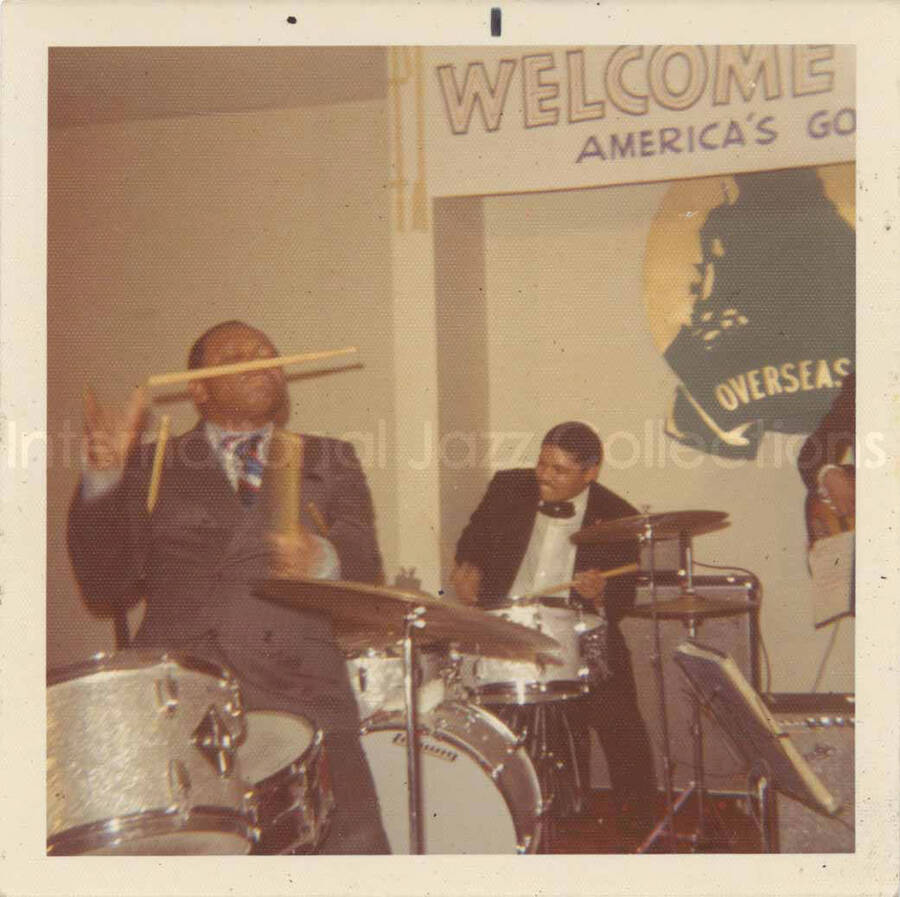 3 1/2 x 3 1/2 inch photograph. Lionel Hampton and band at the Overseas Press Club of America. A banner on the wall reads: Welcome Lionel Hampton, America's Good Will Ambassador of Jazz