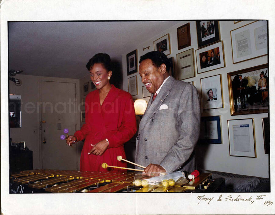 8 3/4 x 13 1/4 inch photograph. Lionel Hampton playing the vibraphone with an unidentified woman, in front of a wall displaying awards, in his apartment. See also LH.III.0859