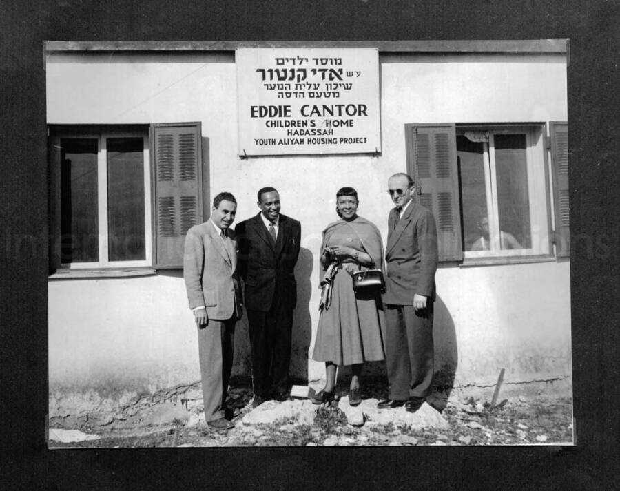 10 x 10 inch photograph. Lionel Hampton in Israel. Gladys and Lionel Hampton with unidentified men in front of a building with a plaque on a wall that reads: Eddie Cantor; Children's Home; Hadassah Youth Aliyah Housing Project