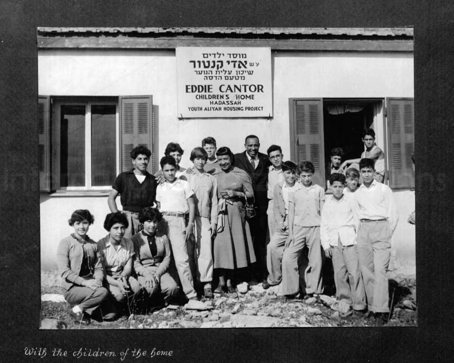 10 x 10 inch photograph. Lionel Hampton in Israel. Gladys and Lionel Hampton with unidentified men in front of a building with a plaque on a wall that reads: Eddie Cantor; Children's Home; Hadassah Youth Aliyah Housing Project. Caption under the photograph reads: With the children of the home
