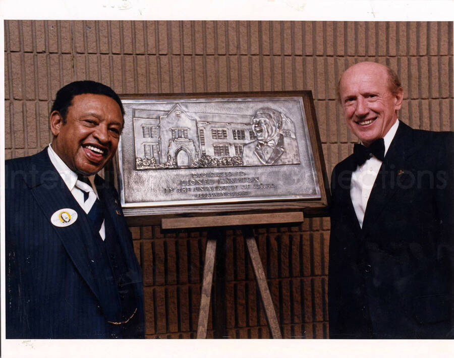 11 x 14 inch photograph. Lionel Hampton with President of the University of Idaho Richard D. Gibb in front of the dedication plaque of the Lionel Hampton School of Music at the University of Idaho, Moscow, ID. This photograph is mounted on cardboard