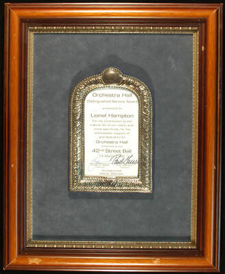 Plaque. 16 1/2"x13 1/2" Wood frame holding a 7 1/2"x5"  silver frame with certificate under glass, in a  and blue velvet background Orchestra Hall Distinguished Service Award presented to Lionel Hampton for his enthusiastic support and dedication to Orchestra Hall, on occasion of the 42nd Street Ball. Frank D. Stella, Chairman and Paul Ganson, President. Detroit, MI, Mar. 14, 1989