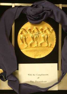 National Medal of Arts presented to Lionel Hampton. 4 inch gold medal designed by sculptor Robert Graham. The medal has a loop on top, from which it is attached by a purple ribbon to an 8 x 7 1/2 x 2 inch display.  On the obverse it reads: National Medal of Arts, and on the reverse: United States of America; Lionel Hampton. Accompanying the medal is a 3/4 inch version of it, on a blue ribbon attached by a pin to a 34 inch long purple ribbon neck tie. The medal is inside a wood box. Also a 4 x 6 inch glass engraved display