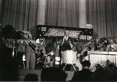 4 x 6 inch photograph. Lionel Hampton performing on the drum with orchestra, which includes guitarist Billy Mackel. A banner on the back of the stage reads: Die Solidaritat Ladt Ein!