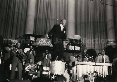 4 x 6 inch photograph. Lionel Hampton performing on the drum with orchestra, which includes guitarist Billy Mackel. A banner on the back of the stage reads: Die Solidaritat Ladt Ein!