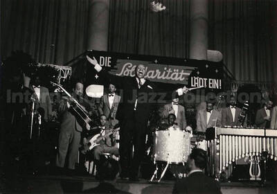 4 x 6 inch photograph. Lionel Hampton performing on the drum with orchestra, which includes guitarist Billy Mackel. A banner on the back of the stage reads: Die Solidaritat Ladt Ein!