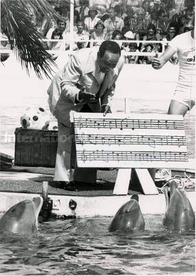 8 x 6 inch photograph. Lionel Hampton holds an enlarged musical score while standing by a pool showing it to dolphins, in front of an audience, at the Marineland. [Antibes, France?]