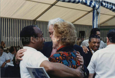 4 x 6 inch photograph. Lionel Hampton, Bill Titone, and unidentified persons under a tent, on the occasion of a ceremony related to wine, [in France]. Lionel Hampton is wearing a red and gold string around his neck. See also the copper ceremonial key in this collection