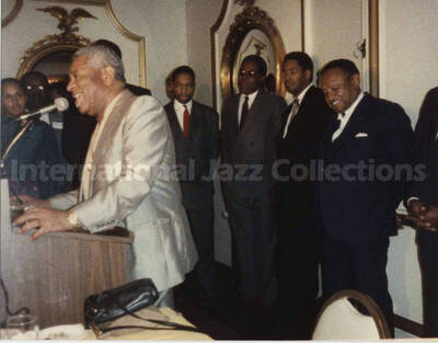 4 x 5 inch photograph. Dizzy Gillespie speaks at a podium observed by unidentified musicians at a reception