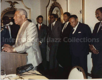 4 x 5 inch photograph. Dizzy Gillespie speaks at a podium observed by unidentified musicians at a reception