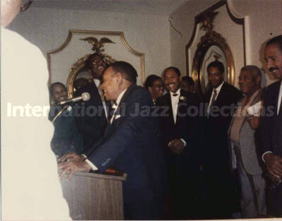 4 x 5 inch photograph. Lionel Hampton speaks at a podium observed by unidentified musicians at a reception
