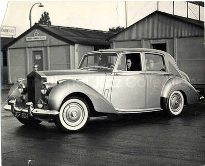 8 x 10 inch photograph. Unidentified persons inside a car parked in front of a building named the Trans-Canada. Handwritten on the back of the photograph: The Silver Pigeon meets