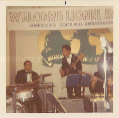 3 1/2 x 3 1/2 inch photograph. Lionel Hampton and band at the Overseas Press Club of America. A banner on the wall reads: Welcome Lionel Hampton, America's Good Will Ambassador of Jazz
