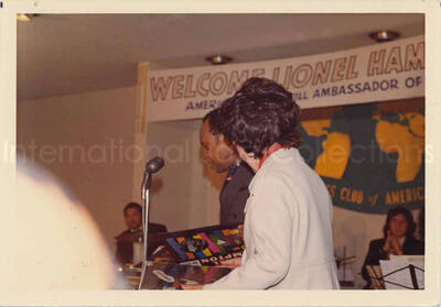 3 1/2 x 5 inch photograph. Lionel Hampton and band at the Overseas Press Club of America. A banner on the wall reads: Welcome Lionel Hampton, America's Good Will Ambassador of Jazz