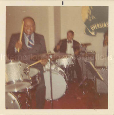 3 1/2 x 3 1/2 inch photograph. Lionel Hampton and band at the Overseas Press Club of America. A banner on the wall reads: Welcome Lionel Hampton, America's Good Will Ambassador of Jazz