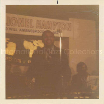 3 1/2 x 3 1/2 inch photograph. Lionel Hampton and band at the Overseas Press Club of America. A banner on the wall reads: Welcome Lionel Hampton, America's Good Will Ambassador of Jazz