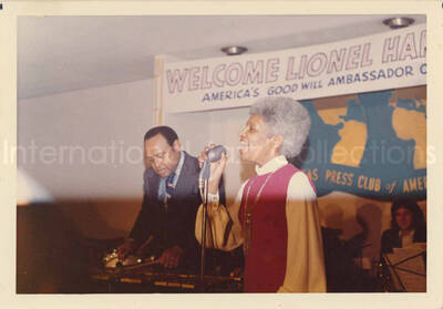 3 1/2 x 5 inch photograph. Lionel Hampton and band at the Overseas Press Club of America. A banner on the wall reads: Welcome Lionel Hampton, America's Good Will Ambassador of Jazz