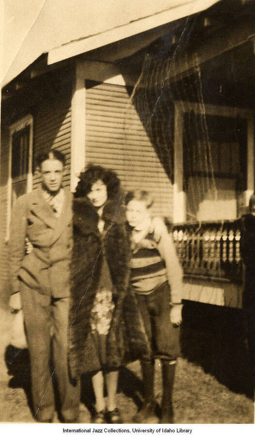 Lee Morse dressed in a fur jacket stands with her arms around a man and child in front of a house.