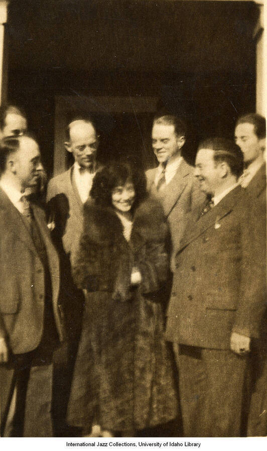 Lee Morse stands in a fur jacket with a crowd of men standing around her on the porch of a house.