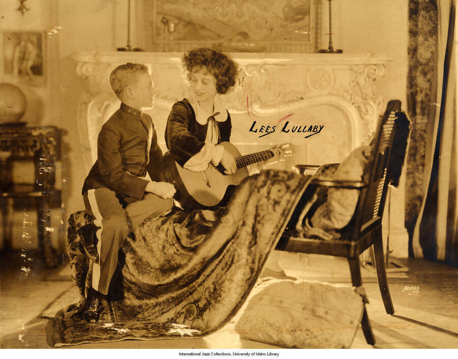 Lee's Lullaby." Lee Morse sits on a chair in front of a fireplace. She holds a guitar and stares at a young child sitting next to her dressed in a uniform.