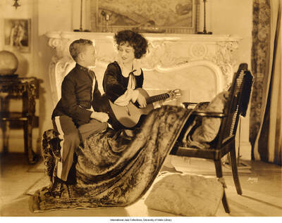 Lee Morse sits on a chair in front of a fireplace. She holds a guitar and gazes at a young child sitting next to her dressed in a uniform.