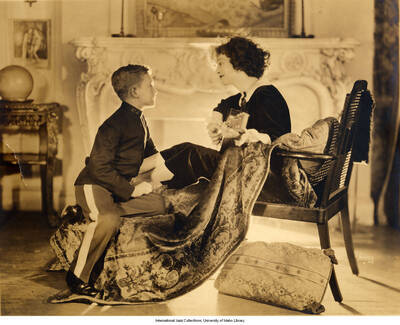 Lee Morse sits on a chair in front of a fireplace. She holds a guitar and gazes at a young child sitting next to her dressed in a uniform.