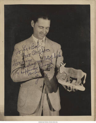 Black and white photograph of a man holding a donkey figurine. Inscription: "To Lee and Bob.  Two Stomp Down Gooduns.  Wishing you lot's of luck + happiness Always  woe Buck woe.  Zeke"