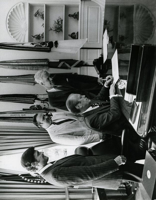 Senator James A. McClure stands with others in the White House Oval Office while President Gerald Ford signs a bill at his desk.