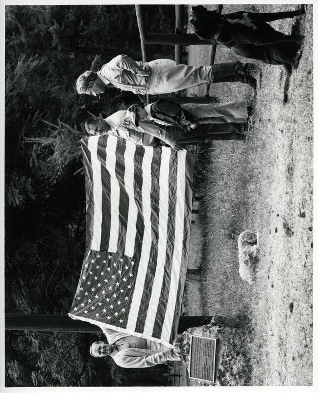 Senator James A. McClure and Senator John Warner stand with unknown others and a dog while presenting a U.S. flag.