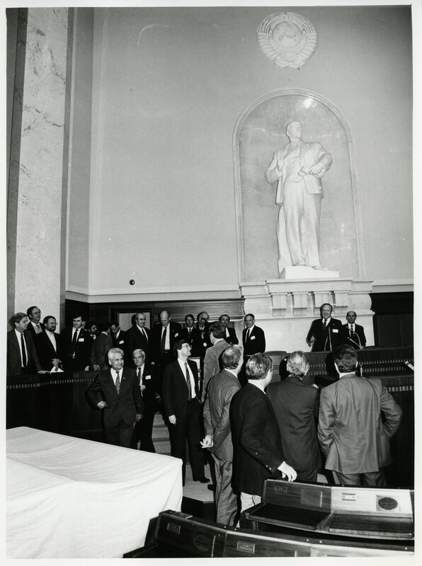 Senator James A. McClure stands at a rostrum before a large statue of Vladimir Lenin as others mill about nearby.