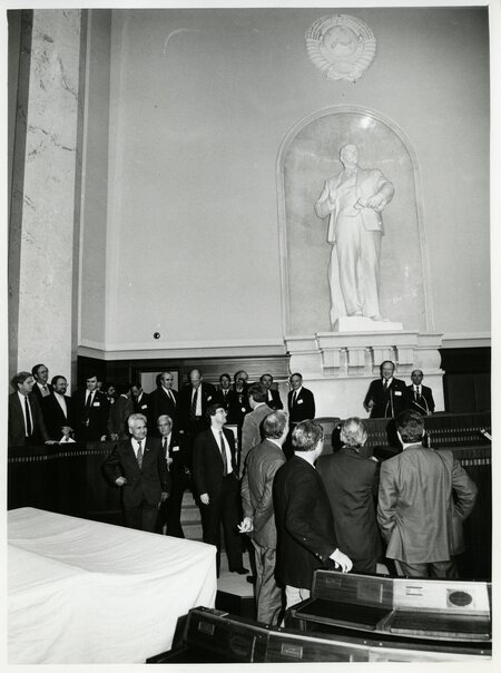 Senator James A. McClure stands at a rostrum before a large statue of Vladimir Lenin as others mill about nearby.