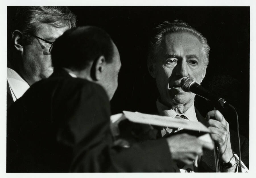 7" x 5" black and white photograph. Leonard Feather speaks into a microphone at the jazz festival. Lionel Hampton and Lynn "Doc" Skinner face him.