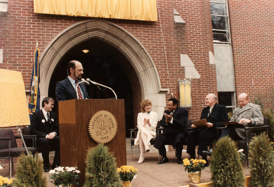 12" x 8" color photograph. School of Music Director Robert Miller speaking at the Lionel Hampton School of Music dedication. Lionel Hampton, Lynn "Doc" Skinner, President Richard D. Gibb, Governor Cecil Andrus, Lynn St. James from Chicago, and an unidentified woman sitting in the background.