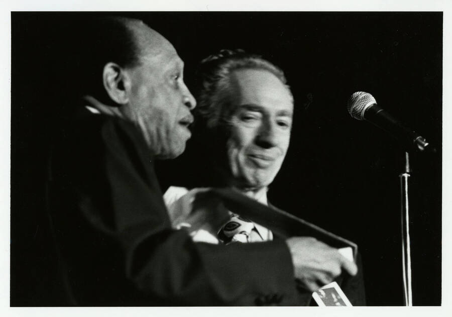 7" x 5" black and white photograph. Lionel Hampton speaks into a microphone while standing next to Leonard Feather. Feather is out of focus in the photograph.