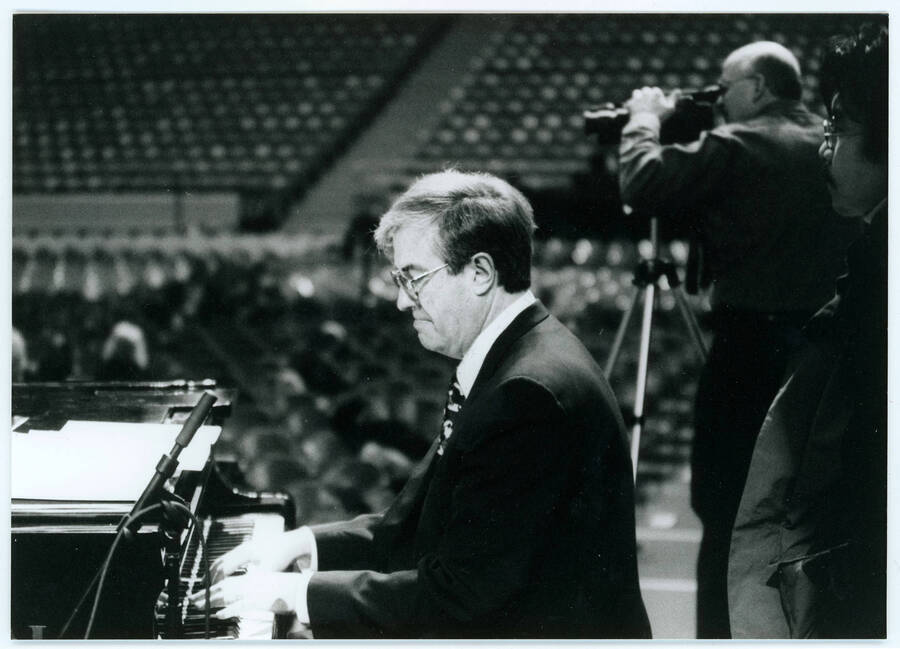 7" x 5 " black and white photograph. Lynn "Doc" Skinner playing piano possibly with Kuni Mikami standing behind him and a camera man in the background. Possibly at a soundcheck for one of the festival concerts.