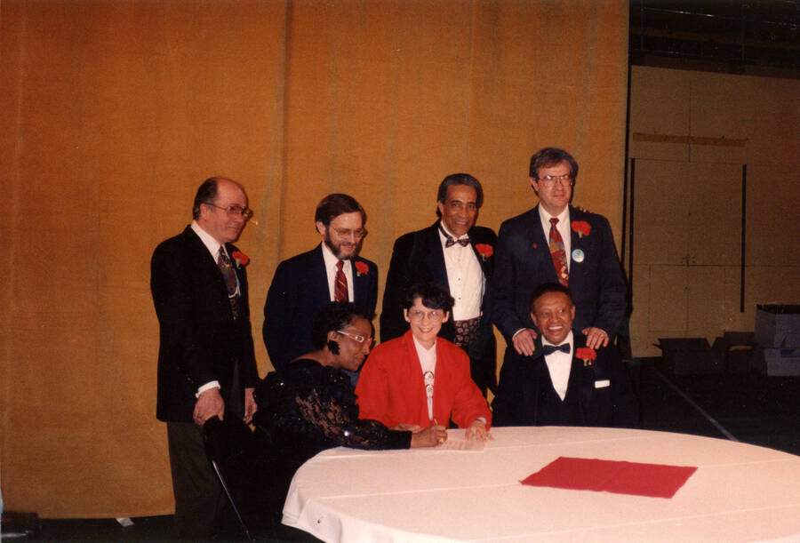 12" x 8" color photograph. President Elizabeth Zinser poses while signing a document. Lionel Hampton, Lynn "Doc" Skinner, School of Music Director Robert Miller, Judge Myron H. Wahls and two unidentified individuals are positioned around her. The photograph was possibly taken at the Red Carnation due to the carnations worn by subjects.