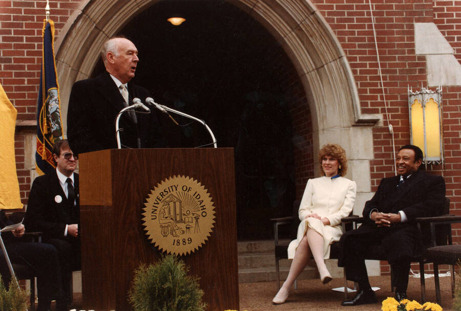 12" x 8" color photograph. Governor Cecil Andrus speaking at the Lionel Hampton School of Music dedication. Lionel Hampton, Lynn "Doc" Skinner, Lynn St. James from Chicago, and an unidentified woman sitting in the background.