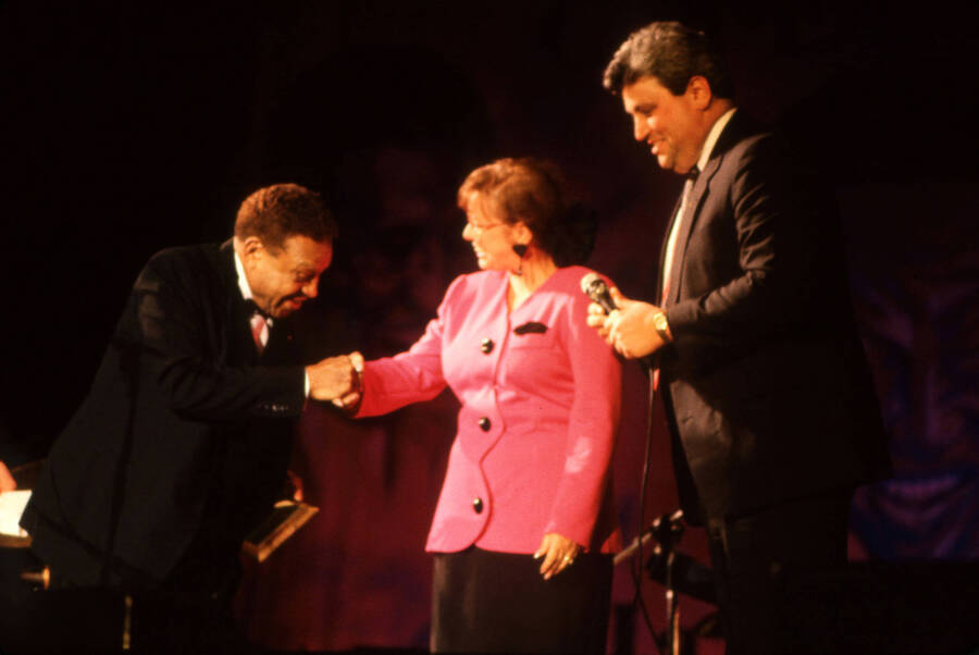 35 mm color slide. An unidentified woman shakes Lionel Hampton's hand on stage  at the Pepsi International World Jazz Night at the 1992 Lionel Hampton-Chevron Jazz Festival. An unidentified man stands next to them holding a microphone.