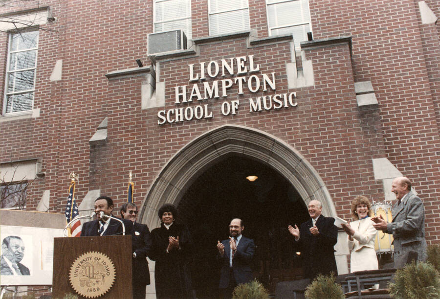 12" x 8" color photograph. Lionel Hampton speaking at the Lionel Hampton School of Music dedication. Lynn "Doc" Skinner, President Richard D. Gibb, Governor Cecil Andrus, School of Music Director Robert Miller, Lynn St. James from Chicago, and an unidentified woman stand clapping in the background.