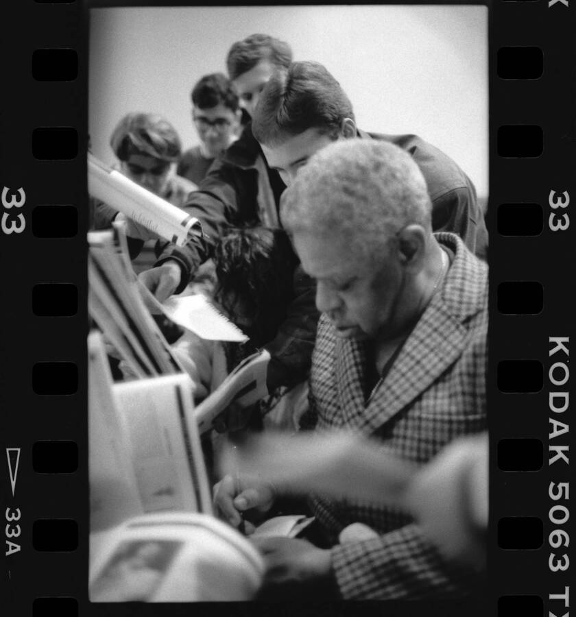 1" x 1.5" black and white print. One of thirty images found on a contact sheet. Dizzy Gillespie signing jazz festival programs for students.
