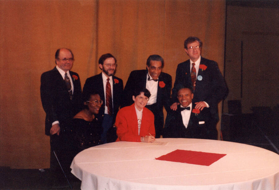 12" x 8" color photograph. Unidentified woman signing a document. Lionel Hampton, Lynn "Doc" Skinner, President Elizabeth Zinser, School of Music Director Robert Miller, Judge Myron H. Wahls and two unidentified individuals are positioned around her. The photograph was possibly taken at the Red Carnation due to the carnations worn by subjects.