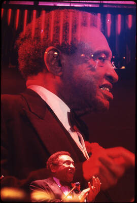 35mm color slide. Lionel Hampton clapping in front of a larger photograph of him.