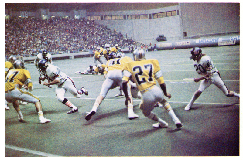 Color photograph of football players in two different styles of football uniforms running on a football field. In the background are stands crowded with people.