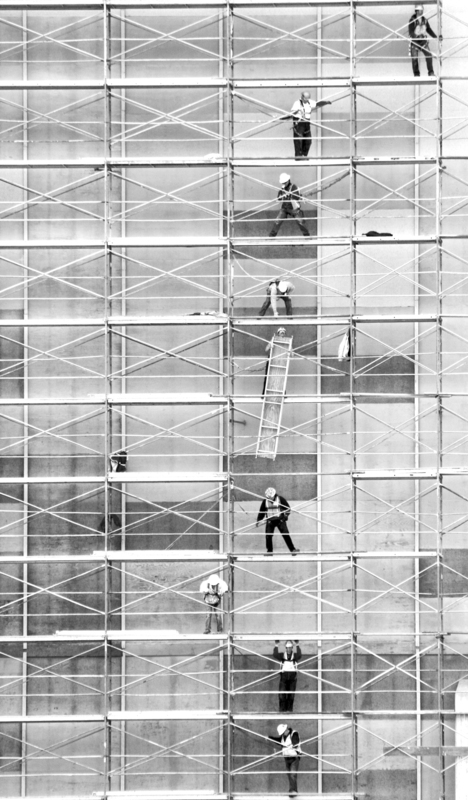 Black and white photograph of people standing on several levels of scaffolding against the side of a building, passing a ladder between each other.