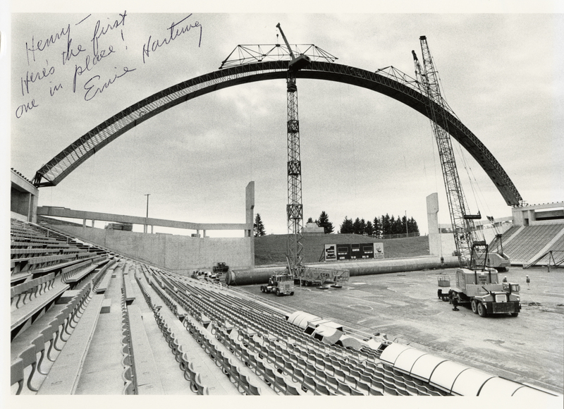 Black and white photograph of an open air stadium. Construction cranes maneuver a wooden arch to attach it to the upper wall of the stadium.